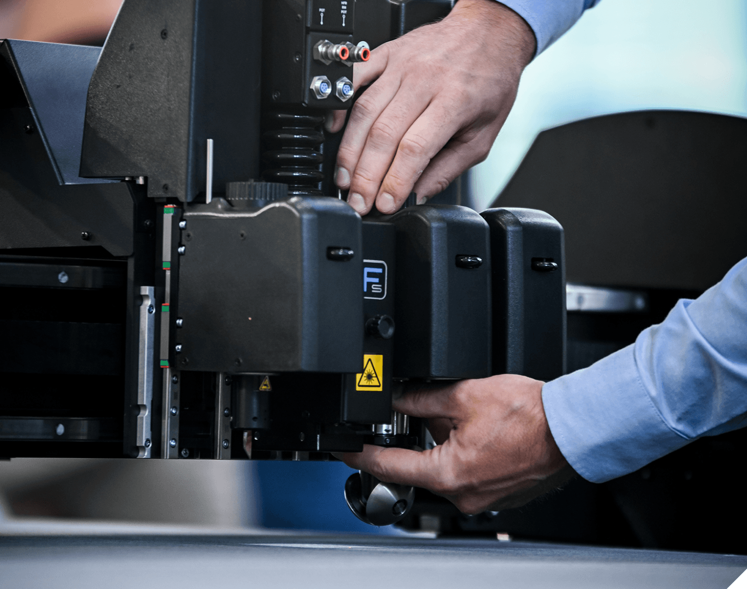 Hands adjusting a machine component with visible warning symbols and connectors, suggesting industrial equipment operation.