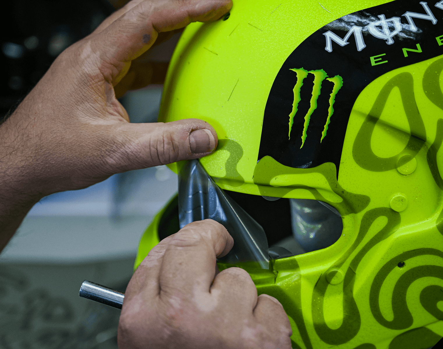 Hands applying a clear film onto a vibrant yellow helmet with a black and green Monster Energy logo and abstract patterns.